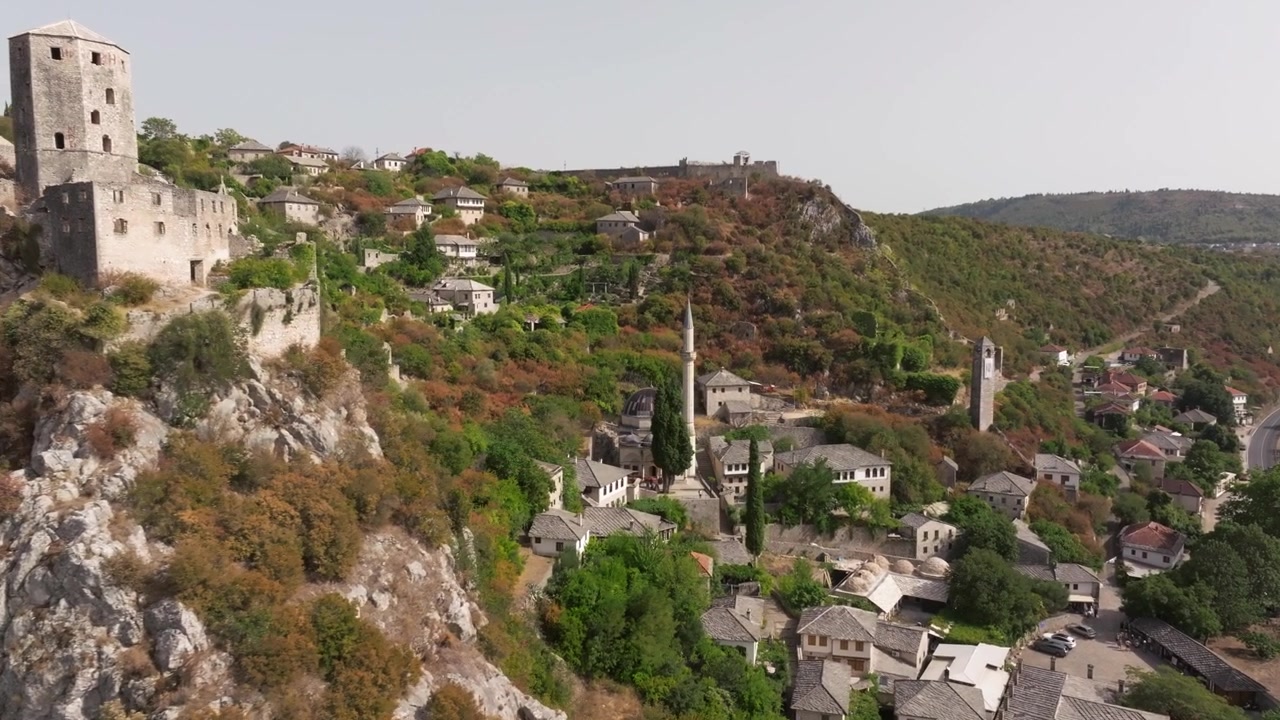 La storica fortezza in pietra di Počitelj e le case in collina si ergono sopra una strada curva e una valle verde durante una ripresa aerea di mezzogiorno