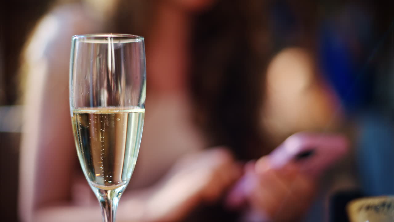Close up of a glass of champagne standing on a table with a woman scrolling on her phone on the background