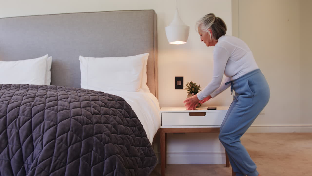 Senior woman holding smartphone, walking in cozy bedroom with smile, at home