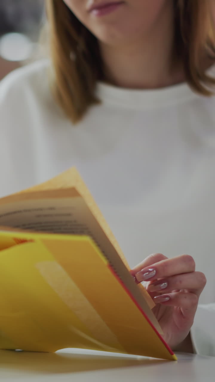 primer plano de una mujer leyendo un libro amarillo en un centro comercial, sosteniendo páginas con una taza de café en la mesa, bolsas de compras de colores visibles, iluminación suave, decoración vibrante y juguetes borrosos en el fondo