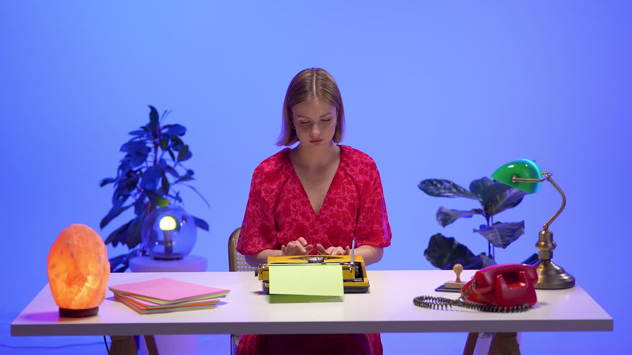 A woman in a red floral dress works on a yellow typewriter in a colorful retro office setup. Surrounded by vintage props, plants, and pastel paper, she focuses quietly in a creative workspace