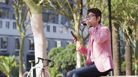 Asiatischer-Mann-Trinkt-Kaffee-Und-Benutzt-Smartphone,-Während-Er-Im-Park-Sitzt