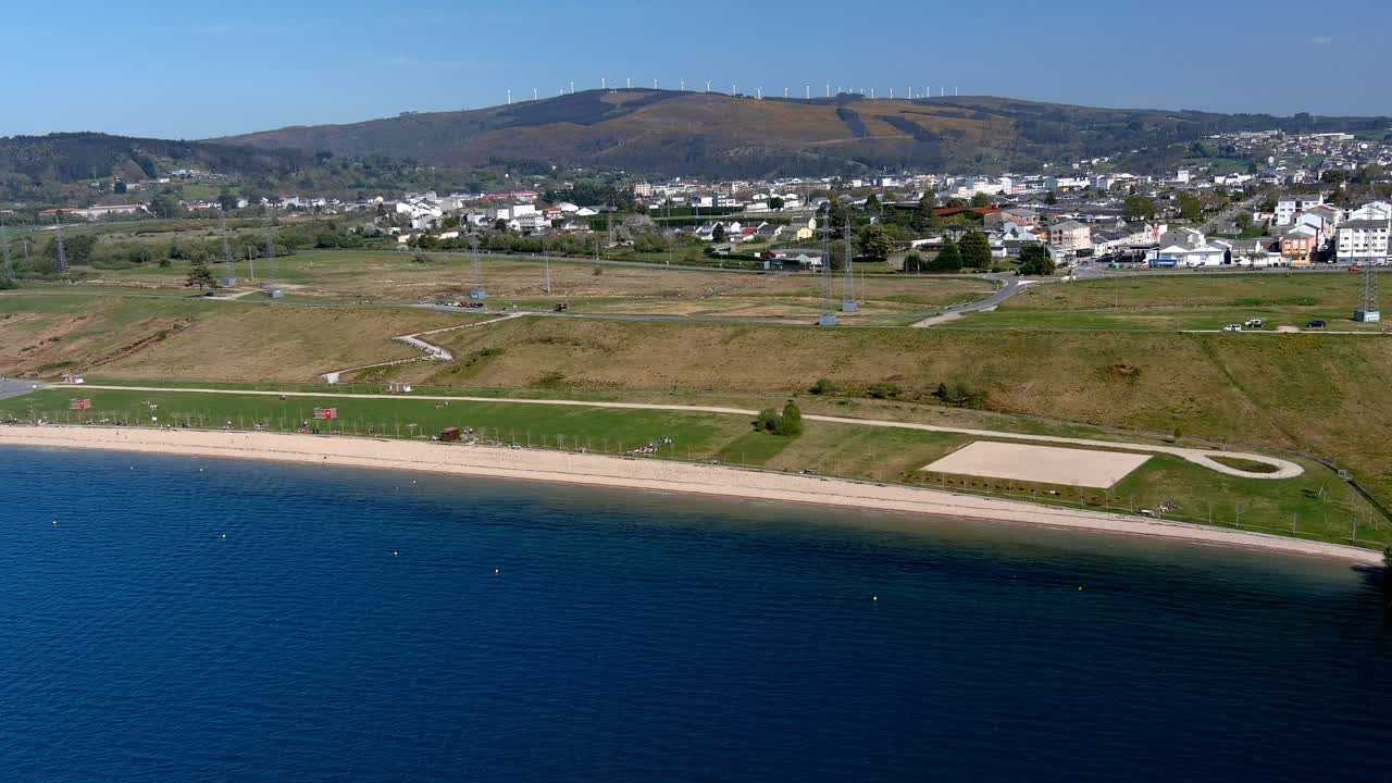 gente en la playa del lago ajardinado con turbinas de viento en el fondo de las montañas, día soleado