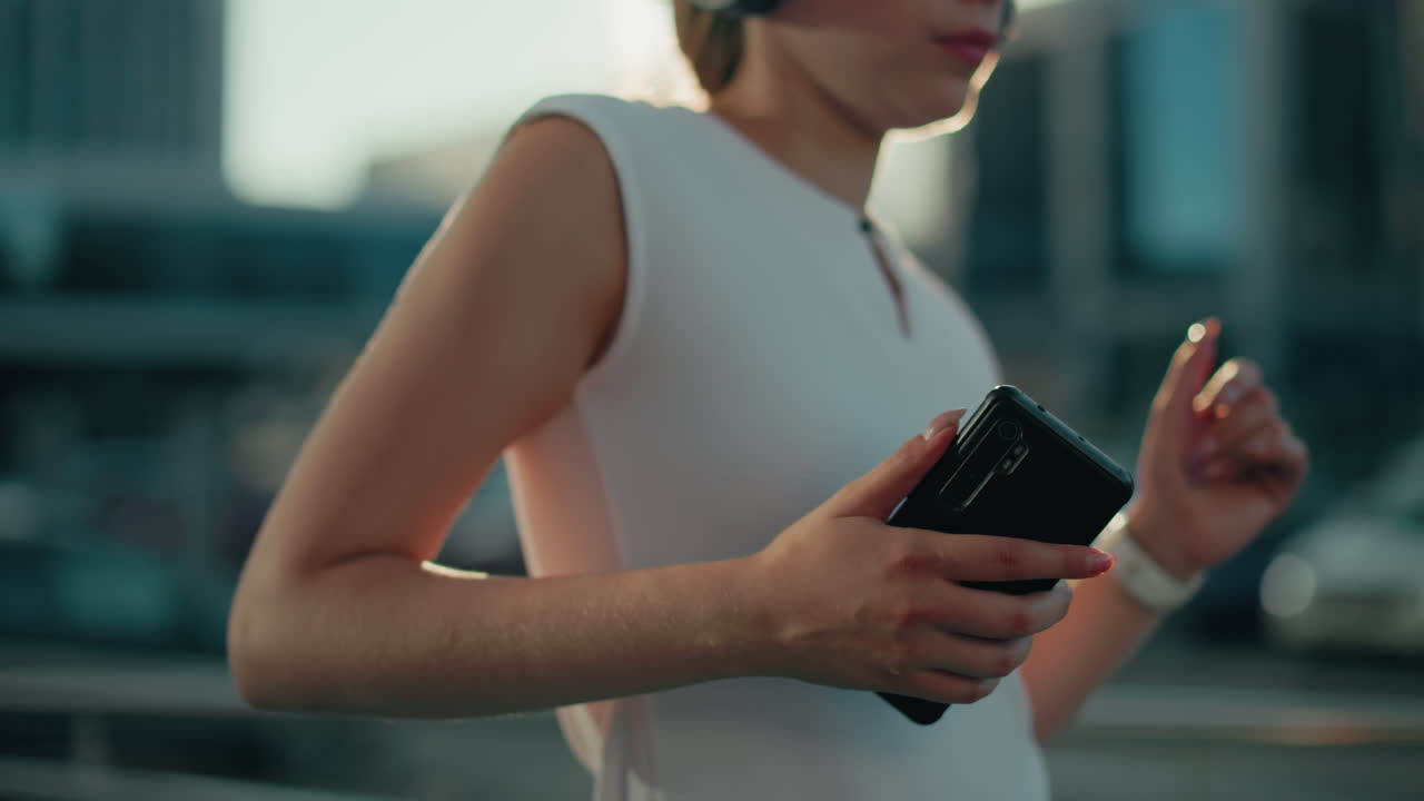 White woman enjoying music with headphones holding phone, sunlight illuminating face and glass building reflecting urban surroundings and hair clipped back