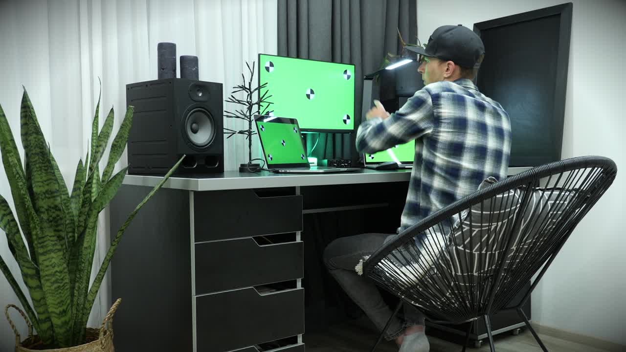 집에서 큰 디스플레이를 가진 컴퓨터에서 일하는 남자, 동료들과 스마트폰으로 이야기하고, 작업 프로세스 작업에 대해 논의합니다. 남자는 녹색 화면의 노트북에서 일합니다.