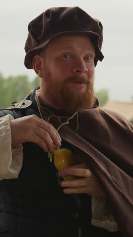 squire with long red beard drinks orange elixir from glass bottle. hero in medieval clothes replenishes strength using magical liquid closeup on blurred background