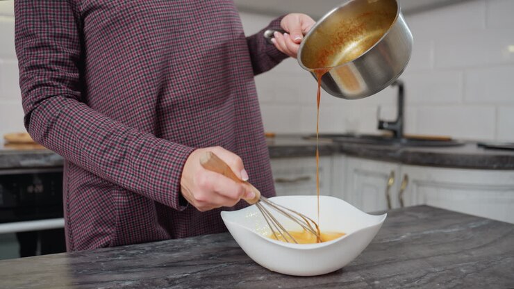 Visuale parziale di una persona che mescola un composto di uova in una ciotola bianca mentre versa una zuppa calda e saporita da una pentola, mani che sbattono e versano attivamente in una cucina moderna con un piano di lavoro in marmo scuro