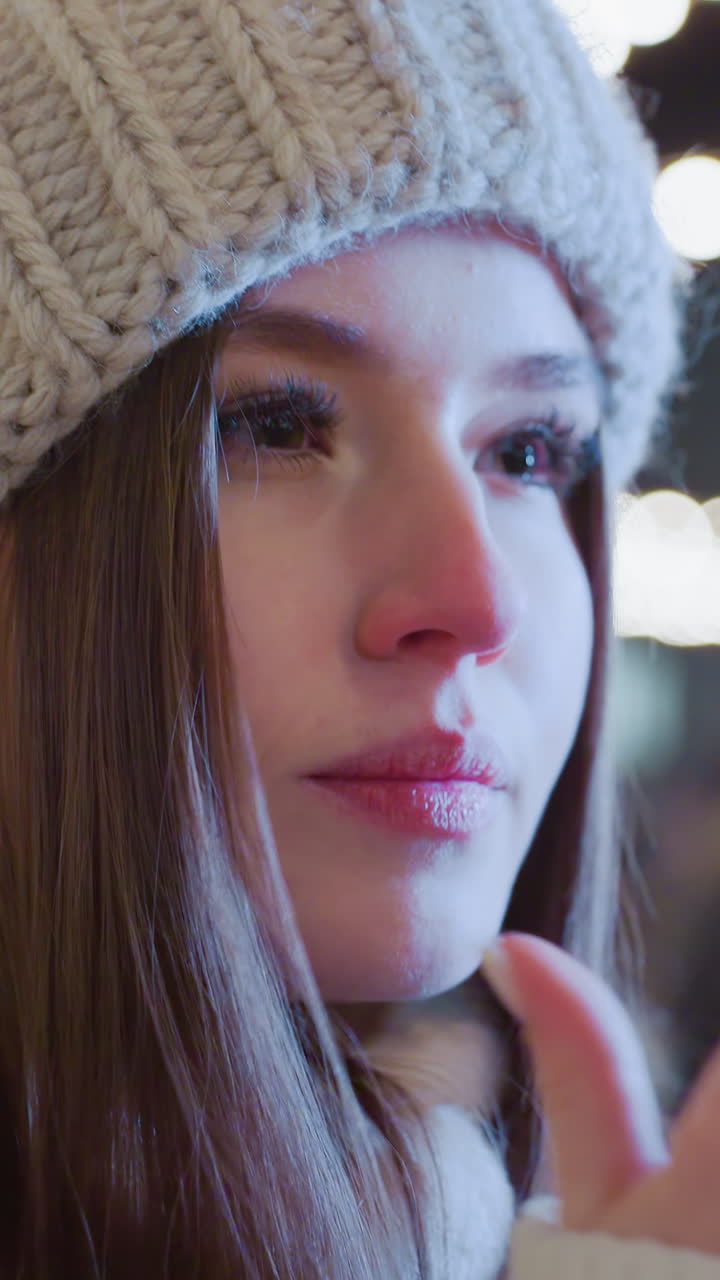 Close-up of young woman in knitted beanie and winter coat, covering her face with hands as she breathes into them for warmth, festive city lights glow in background with people moving around