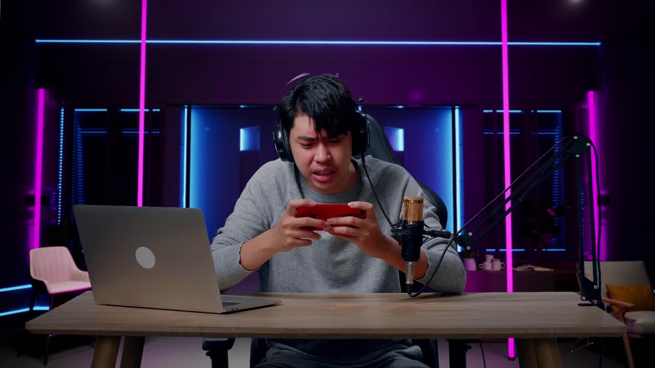 hombre asiático molesto jugador con auriculares y computadora perdiendo juego de teléfono móvil en la habitación iluminada por luces de neón en retro