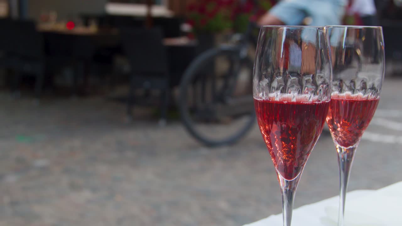 Two crystal glasses of red sparkling wine on table, pedestrians and cyclists passing, soft daylight