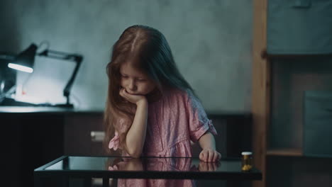 sad girl sits at table with drug bottle in semi-dark room