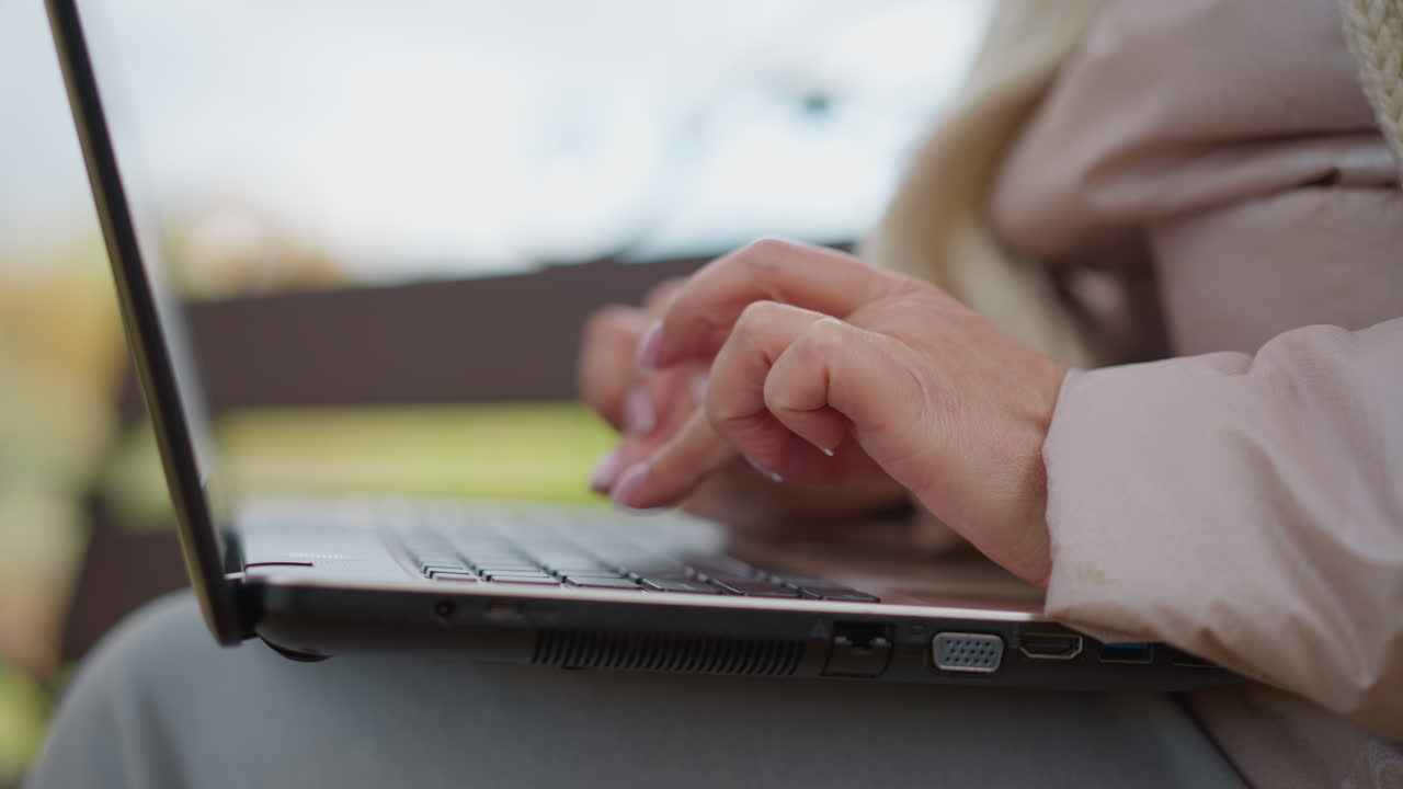 close up side view of woman with painted nails typing on laptop while seated outdoors on bench in autumn park conveying focus and digital work in serene natural setting autumnal background
