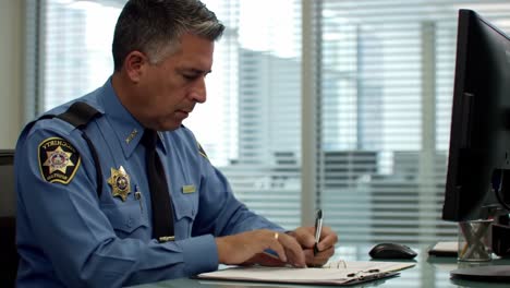 focused law enforcement officer analyzing documents at his desk with pen in hand while interacting with digital technology in modern office environment