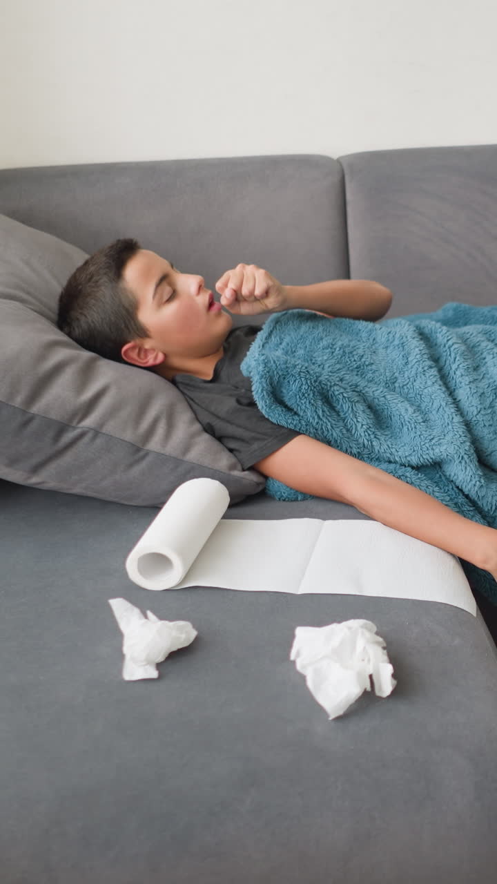 Stanza disordinata con bambino sdraiato su un divano coperto da una coperta, termometro per medicinali e farmaci sul tavolo, bambino che si sistema per stare comodo, scena che indica malattia, riposo e guarigione in un ambiente domestico