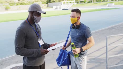 Diverse-male-coach-and-athlete-wearing-face-mask-talking-before-training-session