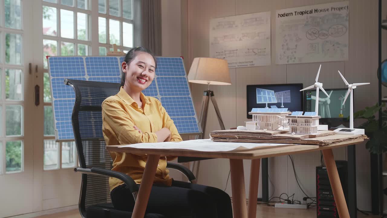 一名亞洲女子交叉雙臂，對著鏡頭微笑，同時正在處理藍圖和附有太陽能板的小屋模型。