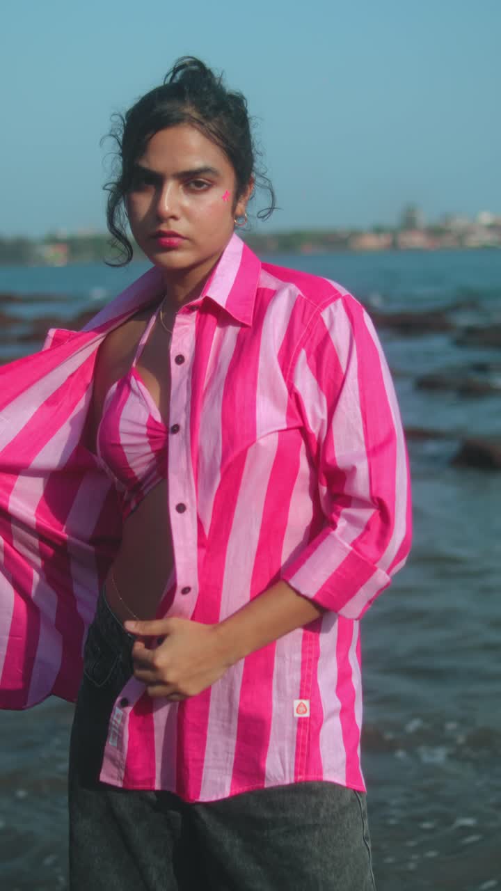 Pink striped shirt, model posing at the beach near the water, with a relaxed vibe