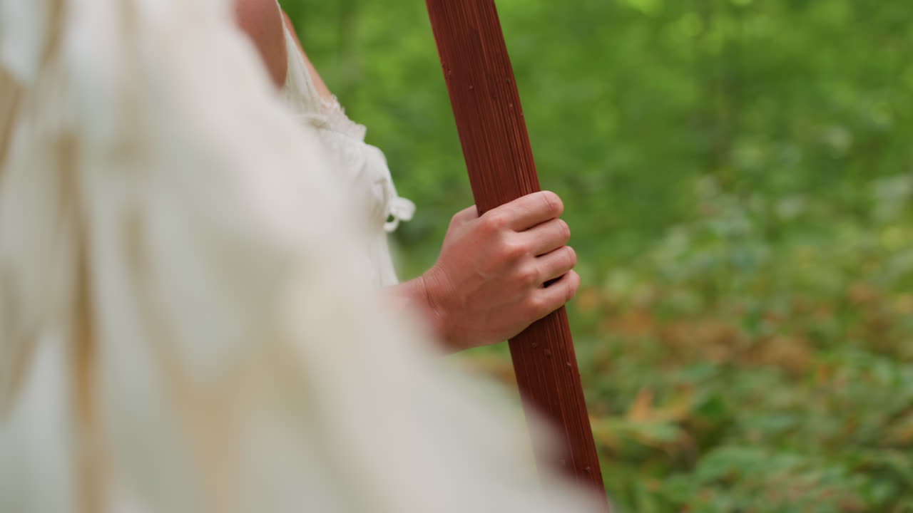 Close up side view of woman dressed in white walking through peaceful forest holding wooden stick in hand surrounded by lush green leaves and soft sunlight symbolizing strength
