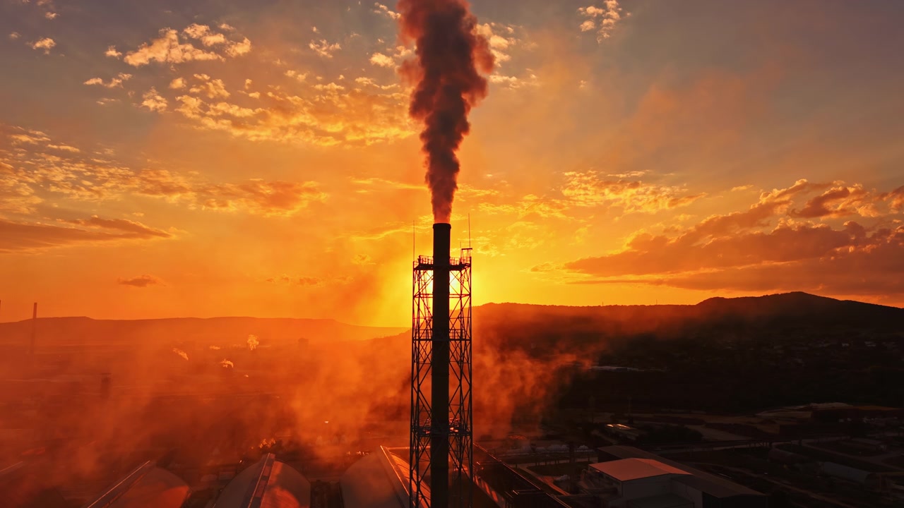 Smoke rises from a factory chimney against a sunset sky with clouds, showing the contrast between industry and nature
