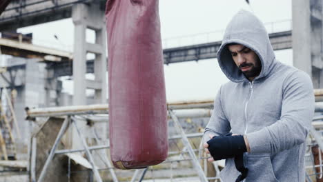 caucasian man wrapping his hand with black cloth and preparing for boxing outdoors an abandoned factory on a cloudy morning