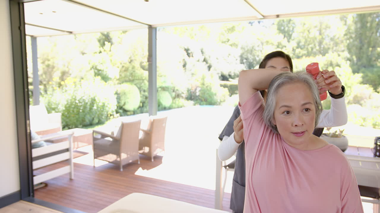 Senior asian woman exercising with dumbbell, assisted by physiotherapist at home