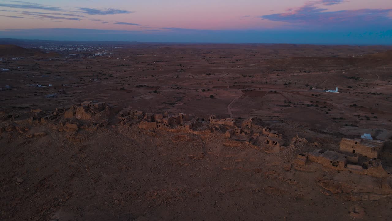 Aerial view of ancient Ksar Jarrat ruins at sunset, overlooking a Tunisian town. Brown, earthy tones dominate the landscape.