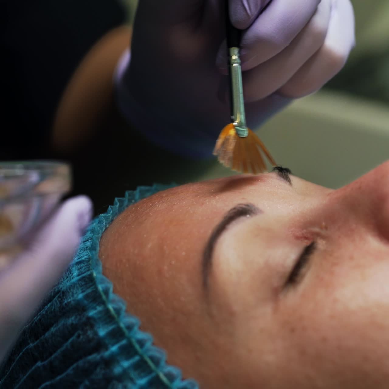 Cosmetologist covers the face of Caucasian woman with lotion. Beautician uses wide brush in work. Extreme close up