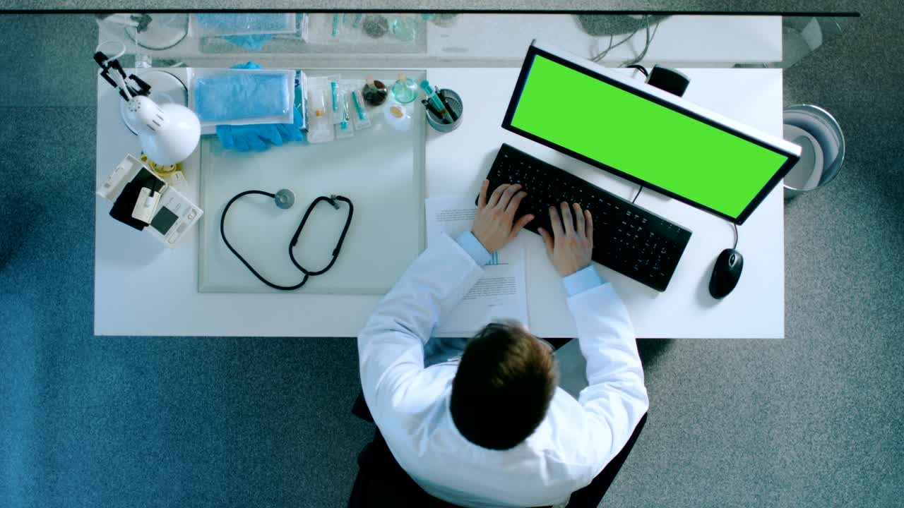 vista superior de un médico sentado en su escritorio y escribiendo en una computadora con pantalla verde. él canta documento de papel.