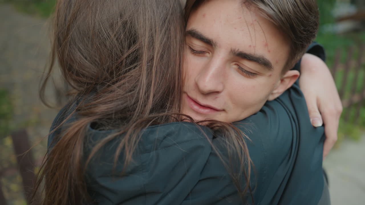 Adolescente bianco riceve un tenero abbraccio su un sentiero vicino a una panchina e del fogliame, occhi chiusi in segno di sollievo, momento intimo di guarigione, implicito conforto da parte di un fratello/sorella o di un counselor, toni caldi, focus cinematografico ridotto