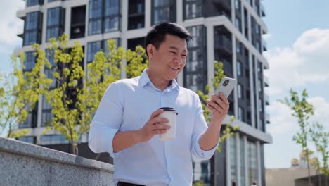 exited handsome asian businessman looking at smartphone drinking coffee outdoor. successful chinese business man reading good news on mobile phone. successful deal, startup bet. finance, triumph.