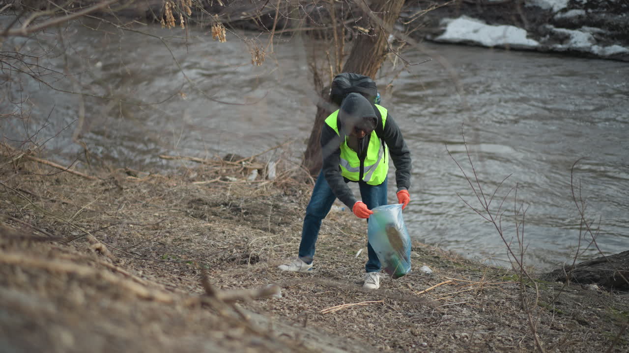 Volunteer in reflective vest, hoodie, and gloves collects plastic bottle from ground near riverbank, placing waste into blue bag during community cleanup to protect environment