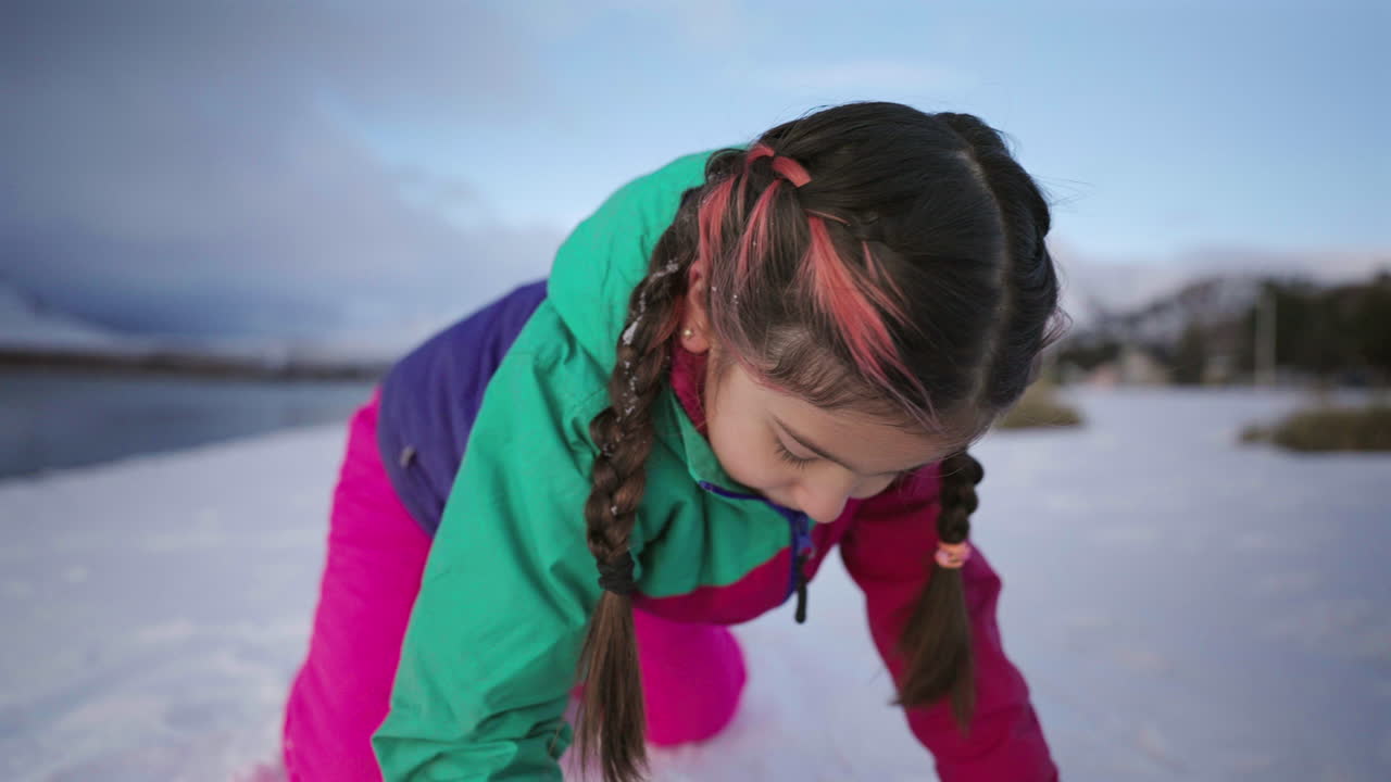 Playful child in colorful jacket making snowball at lakeside winter landscape
