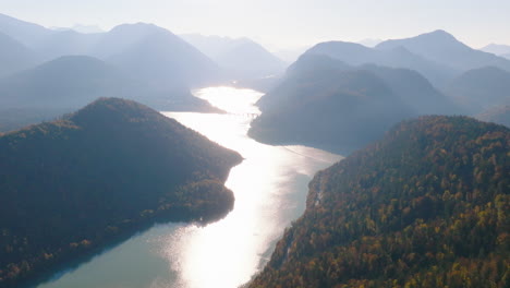 Aerial-view-orbiting-winding-shimmering-Sylvenstein-lake-hazy-Bavaria-mountain-forest-hills