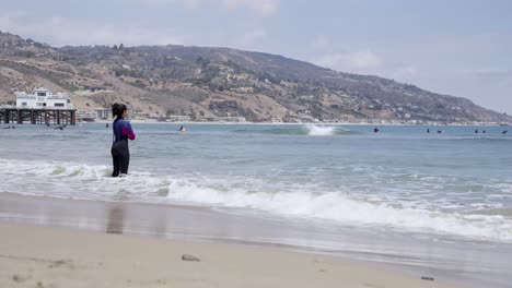 woman surfer standing on the edge of the beach looking after other surfers