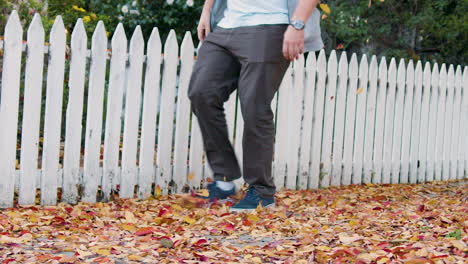 man skips through colorful autumn leaves on sidewalk in front of white picket fence