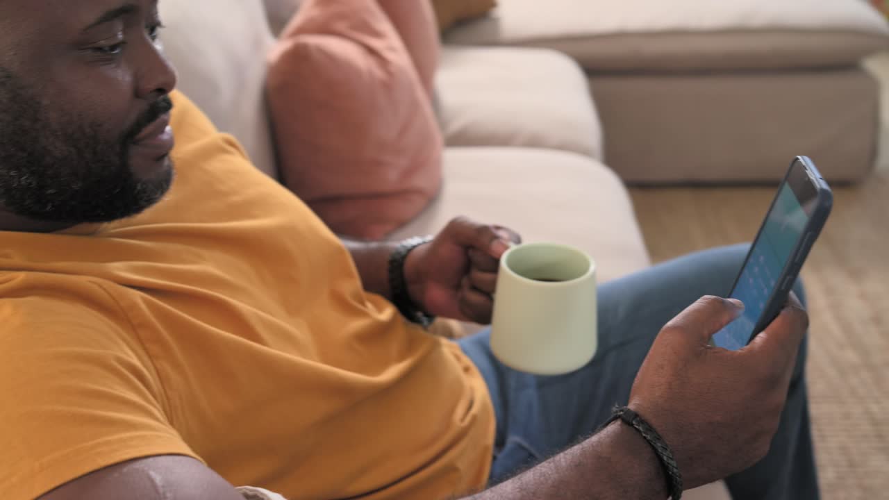 Uomo afroamericano di mezza età con maglietta arancione sdraiato sul divano di casa, che controlla il telefono con un'app e sorseggia da una tazza.