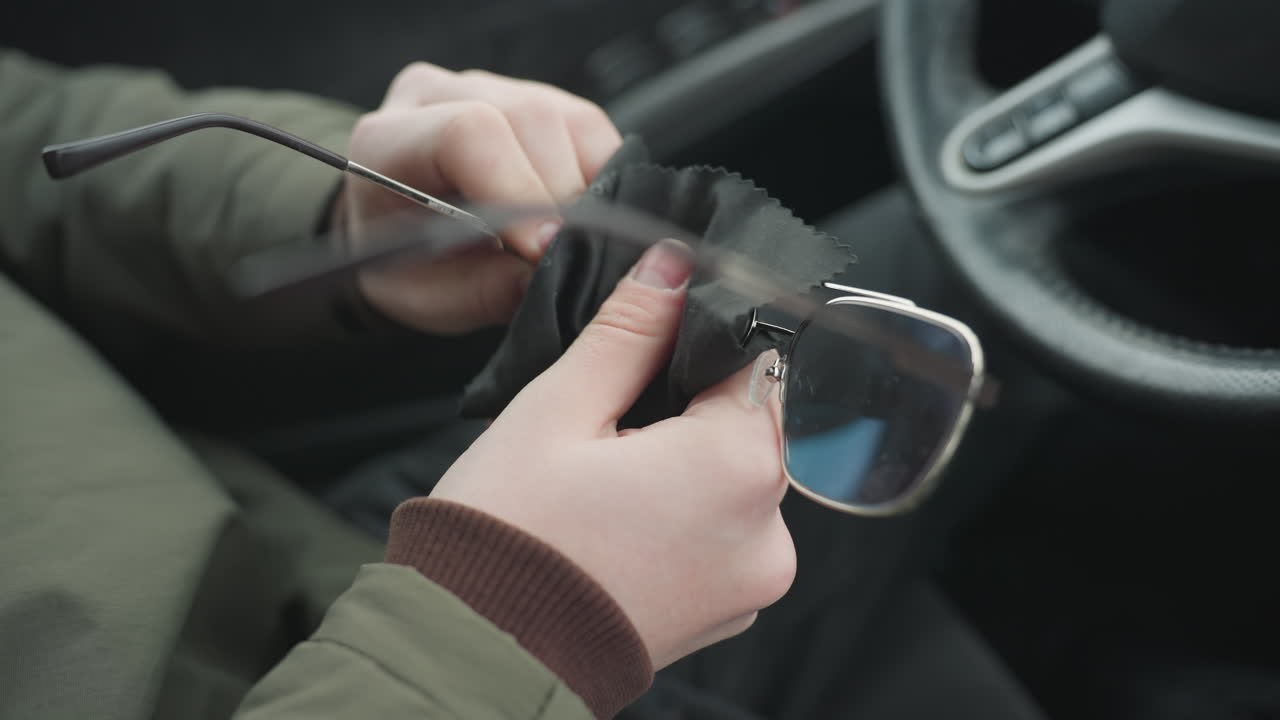 Partial driver view cleaning glasses lens with soft cloth inside car cabin showing hands wiping spectacles near steering wheel under natural daylight capturing maintenance action in modern interior