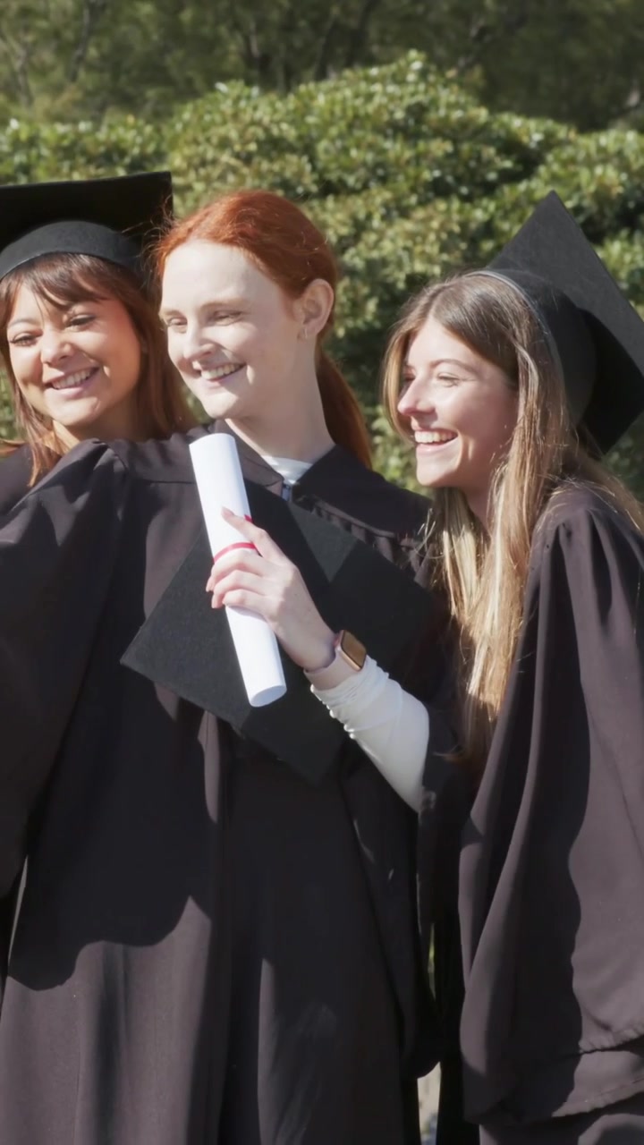 Video verticale: Laureati in toga e tocco che festeggiano, alzano i diplomi e controllano il telefono nel campus.