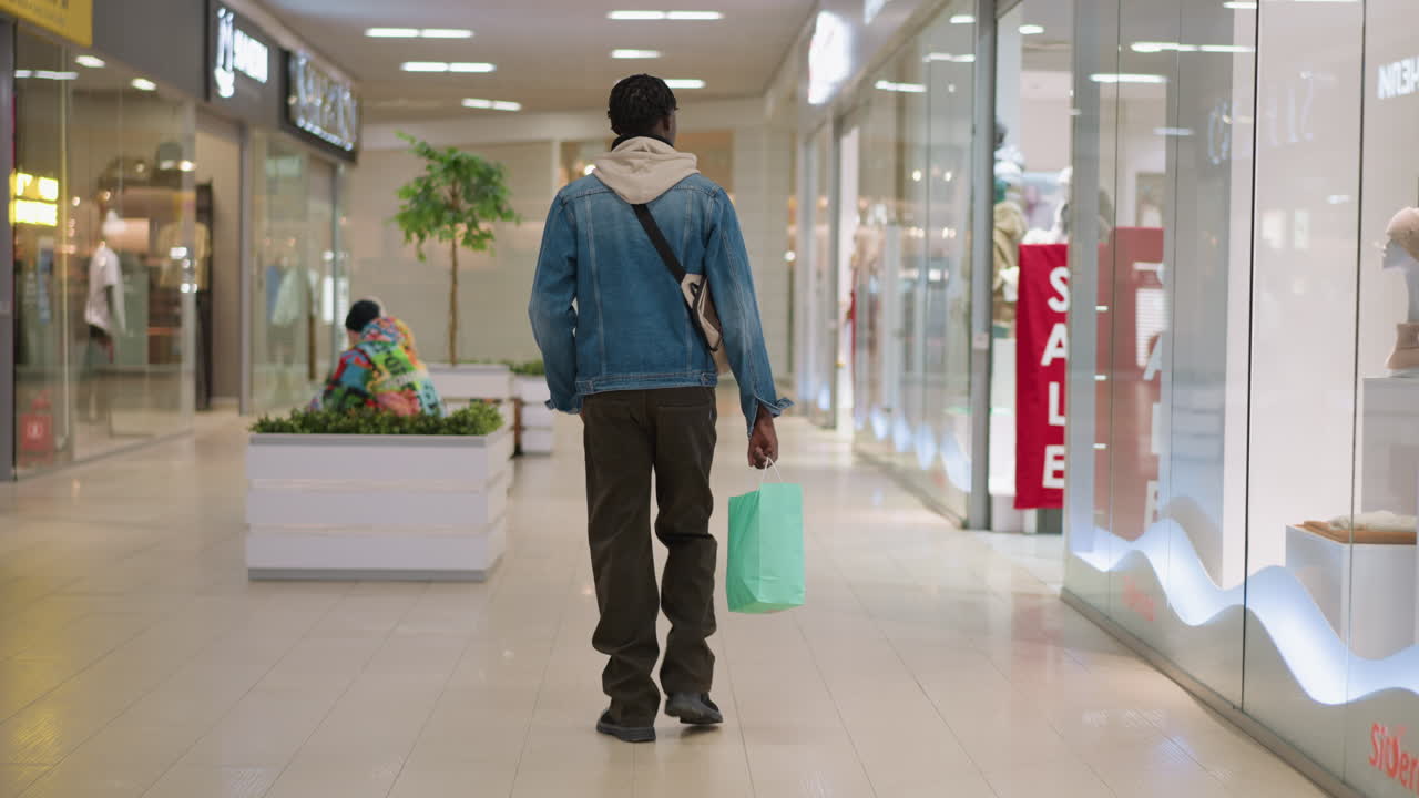 male shopper walking away down bright mall corridor carrying green shopping bag over shoulder wearing denim jacket and crossbody bag passing storefronts and seating planter area interior retail center