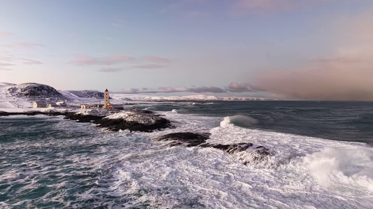 Aerial drone footage ofKjølnes or Kjolnes Lighthouse on a snow covered cliff that is in between wavy ocean sea water during winter time in Norway daytime. Mountains in the back. Lighthouse is white.