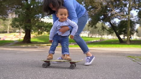 Little-boy-standing-on-skateboard,-rolling-with-help-of-mother