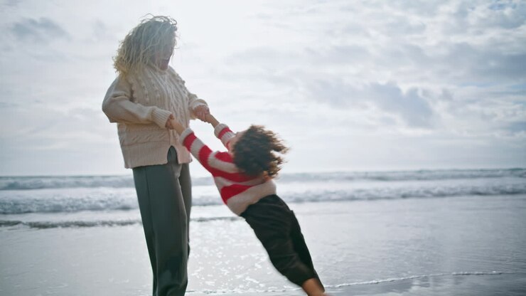 Lachender Elternteil dreht Kind am frühlingshaften Strand. Fröhlicher Junge mit Locken hat Spaß.