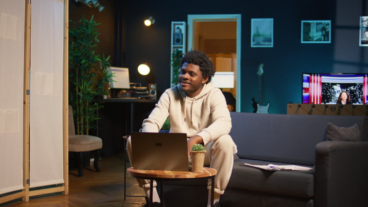 Young male freelancer managing schedules and notes on his laptop while seated in a contemporary living room. Inspiring man looks at pc for a productive teleworking session. Camera B.