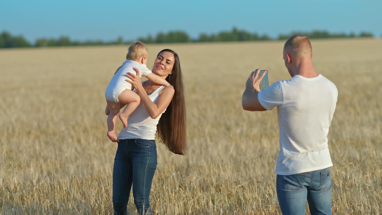 молодой человек снимает видео на свой телефон, когда красивая женщина кружит ребенка. фотосессия на пшеничном поле, блогер делает видео. семейное видео для памяти. 4k, prores
