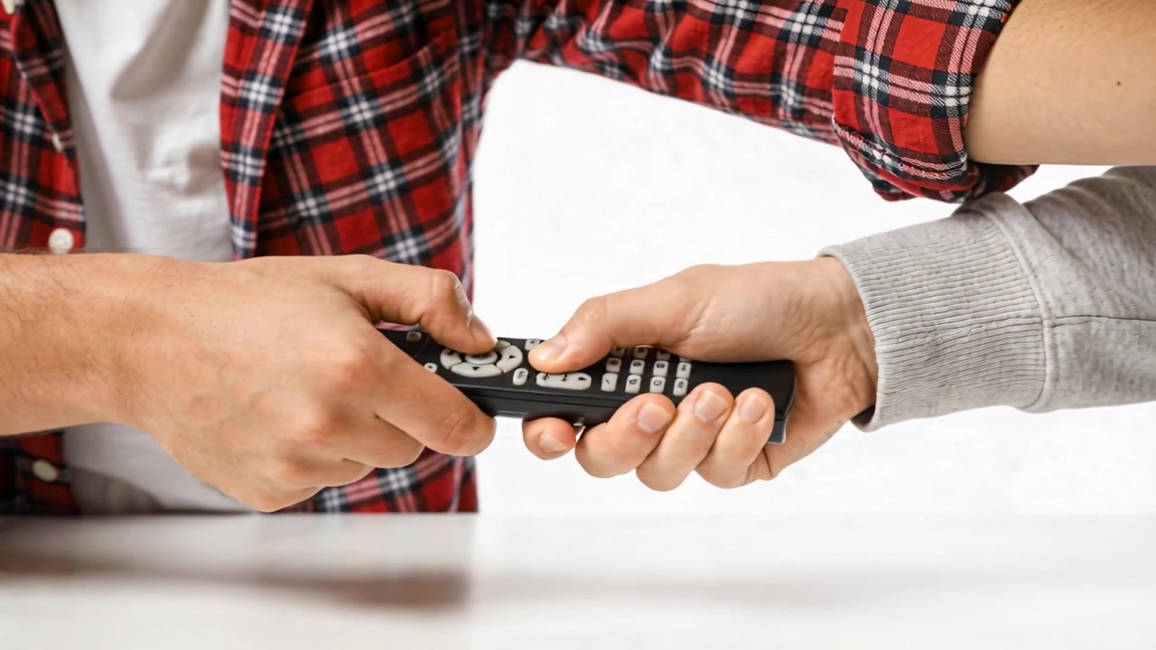 Reaching left in plaid shirt pressing center button while right hand holding remote on white table