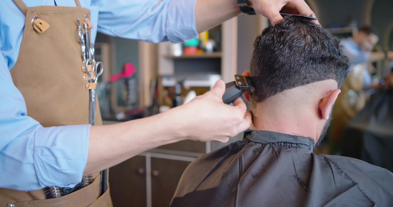 Barbeiro afro-americano de avental marrom começando um corte degradê em um cliente na barbearia com máquina de cortar cabelo, esfumando o cabelo.