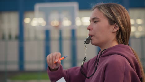 side view of coach with whistle in her mouth directing player on what to do, with blurred background featuring building and sports court, showing focus and instruction during practice