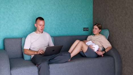 caucasian man is focused on his laptop. wife sits beside on the sofa holding an infant baby.