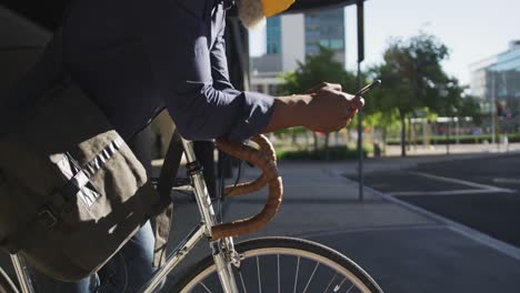 Afroamerikanischer-älterer-Mann-Mit-Gesichtsmaske-Und-Smartphone,-Während-Er-Sich-Auf-Seinem-Fahrrad-Auf-Der-Rechten-Seite-Lehnt