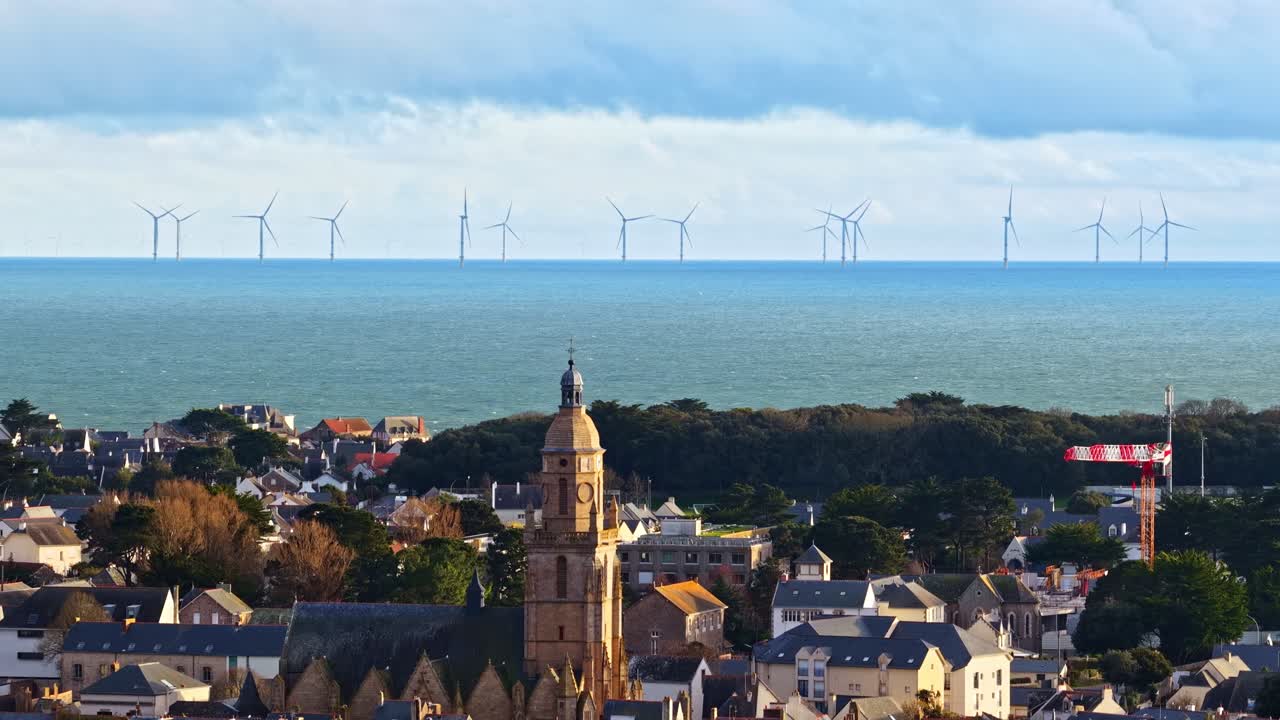 En kystlandsby med et fremtredende kirketårn har utsikt over havet, der vindturbiner preger horisonten. Ideelt for prosjekter om fornybar energi eller kystsamfunn.